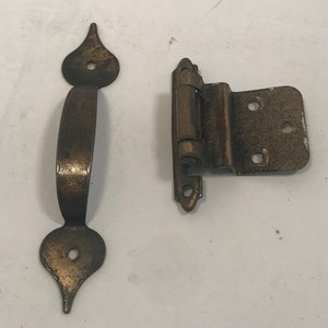 May include: Aged brass cabinet hardware set, including a decorative handle with a curved grip and two ornate end pieces, and a matching hinge. The hardware has a vintage aesthetic, with visible wear and a warm, metallic finish.