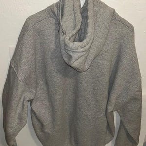May include: A gray hooded sweatshirt with a drawstring hood. The back of the sweatshirt is visible, showing the hood and the fabric's texture. The sweatshirt is hanging on a white hanger.