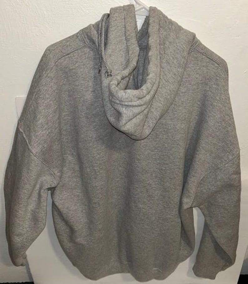 May include: A gray hooded sweatshirt with a drawstring hood. The back of the sweatshirt is visible, showing the hood and the fabric's texture. The sweatshirt is hanging on a white hanger.