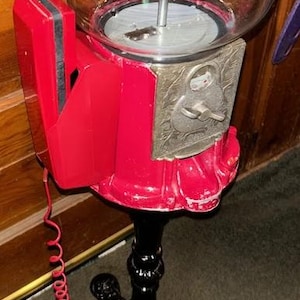 May include: A vintage red gumball machine with a clear glass globe. The machine has a coin slot and a black, ornate base. A red coiled cord and a red plug are attached to the machine.
