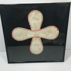 May include: A framed artwork featuring a white baseball stitched together in the shape of a four-pointed star. The baseball is framed against a black background.