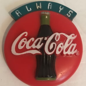 May include: A red and green round button with the words "Always" in a green arch above a white Coca-Cola logo and a brown bottle.