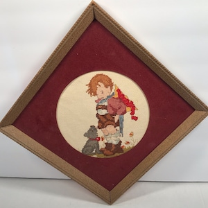 May include: A framed cross-stitch embroidery depicting a young boy with a red backpack and a small dog. The embroidery is set against a cream-colored background and framed with a brown wood frame.
