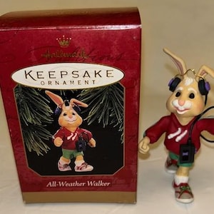 May include: A Hallmark Keepsake ornament featuring a running bunny wearing headphones and a red sweatshirt. The ornament is accompanied by a red box with the words "Keepsake Ornament" and "All-Weather Walker".