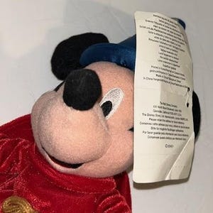 May include: A plush Mickey Mouse toy with a red velvet outfit, black ears, and a blue hat. The toy has a tag attached with printed text. The toy is a Disney product.