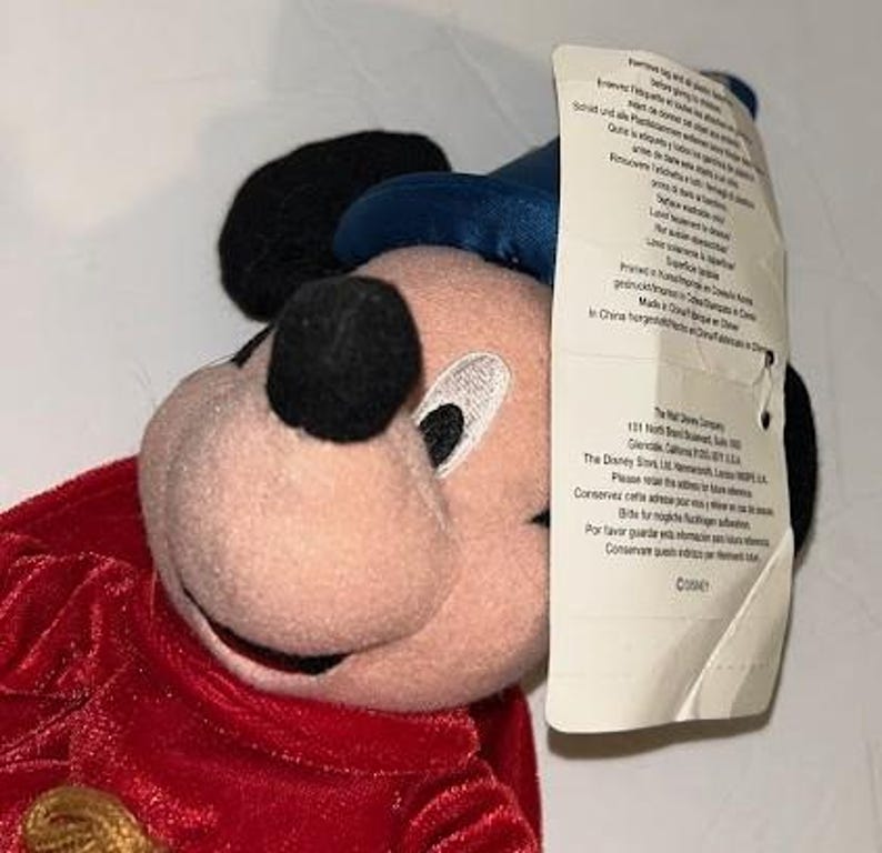 May include: A plush Mickey Mouse toy with a red velvet outfit, black ears, and a blue hat. The toy has a tag attached with printed text. The toy is a Disney product.
