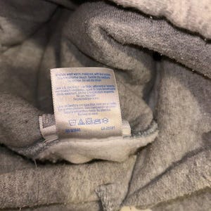 May include: Close-up of a gray sweatshirt with a white tag. The tag includes washing instructions and the text "CA 25181". The sweatshirt appears to be made of a soft, fleecy material.