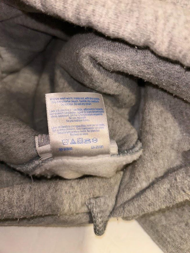 May include: Close-up of a gray sweatshirt with a white tag. The tag includes washing instructions and the text "CA 25181". The sweatshirt appears to be made of a soft, fleecy material.