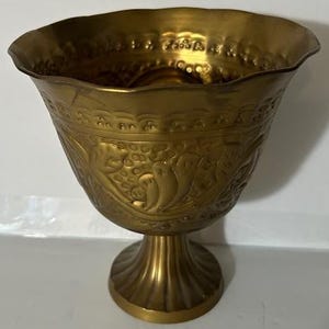May include: A gold metal vase with an ornate design. The vase has a wide, flared top and a round base.