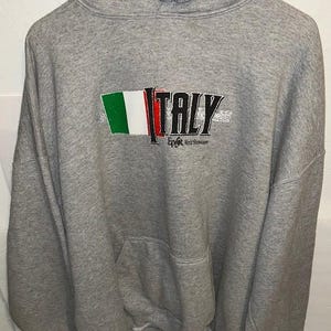 May include: Gray hooded sweatshirt with a front pocket and drawstring. The sweatshirt features the word "ITALY" in black letters with the Italian flag colors and the text "Epcot World Showcase".