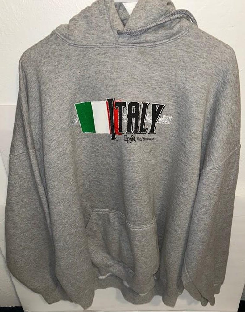 May include: Gray hooded sweatshirt with a front pocket and drawstring. The sweatshirt features the word "ITALY" in black letters with the Italian flag colors and the text "Epcot World Showcase".