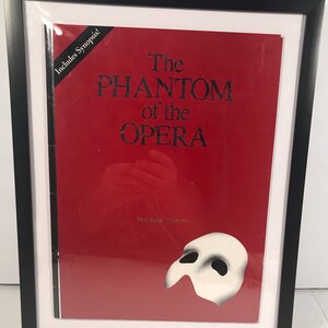 May include: A framed red program for the musical "The Phantom of the Opera" with the title and a white mask in the lower right corner. The program includes a synopsis.