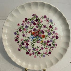 May include: A white ceramic plate with a scalloped edge. The plate features a floral design in the center, with a wreath of purple, pink, and orange flowers. A blue ribbon is tied around the flowers. The plate is supported by a gold-colored stand.
