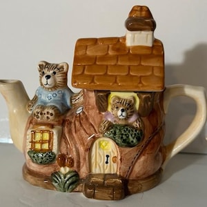 May include: A ceramic teapot shaped like a house with a brown roof and a chimney. Two teddy bears are sitting on the roof and a third teddy bear is peeking out of a window. The teapot has a brown handle and a spout.