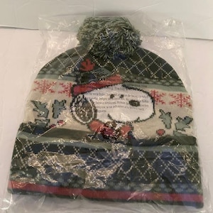 May include: A winter hat with a green pom-pom and a pattern featuring Snoopy. The hat is primarily dark green with a diamond pattern, and a band of red and white near the bottom. The hat is packaged in a clear plastic bag.