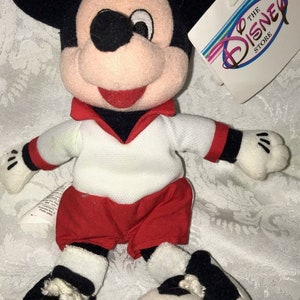 May include: A Mickey Mouse plush toy wearing a white and red soccer uniform, with a black and white soccer ball. The toy has black ears, a pink face, and a red collar. A Disney Store tag is attached.