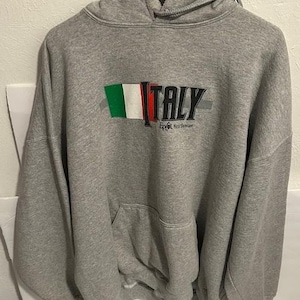 May include: A gray hooded sweatshirt with a front pocket and a drawstring hood. The word "ITALY" is printed across the chest in black, with the Italian flag colors above the word. Below the word "ITALY" is the text "Epcot World Showcase."