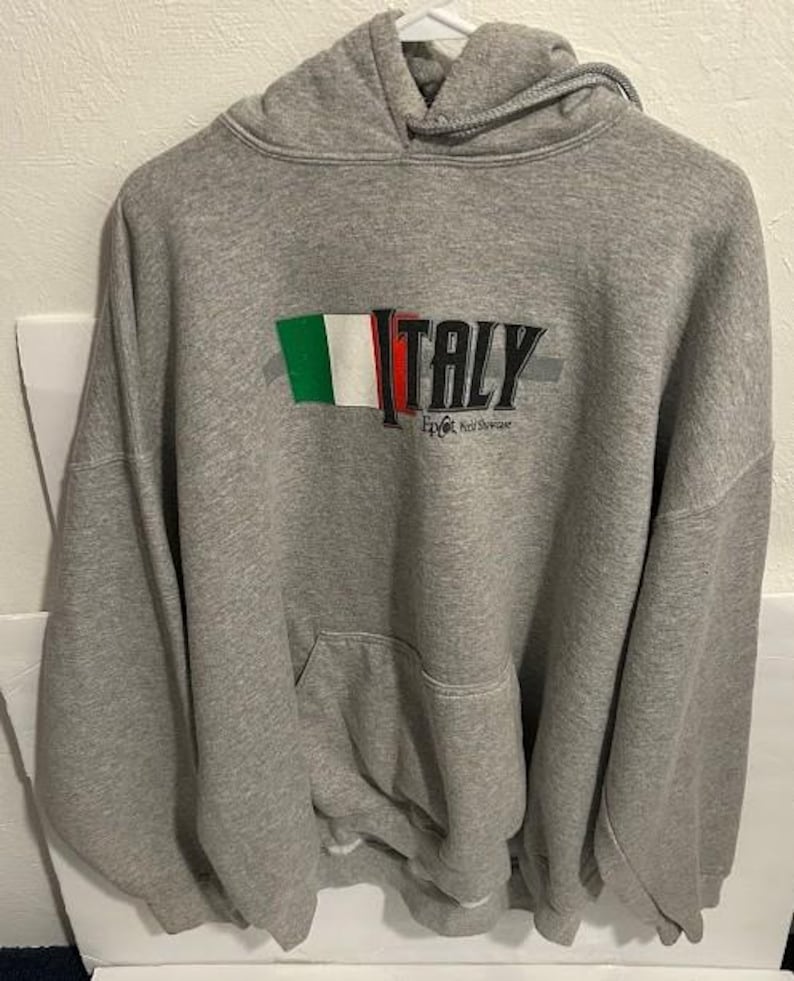 May include: A gray hooded sweatshirt with a front pocket and a drawstring hood. The word "ITALY" is printed across the chest in black, with the Italian flag colors above the word. Below the word "ITALY" is the text "Epcot World Showcase."