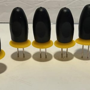 May include: A collection of eight black corn holders with yellow bases and silver prongs. The holders are oval-shaped and designed to securely grip corn on the cob. They are arranged in a slightly curved line.