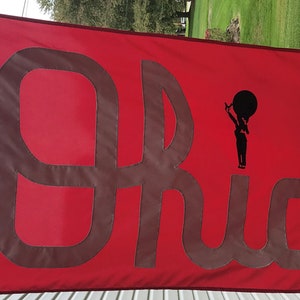 May include: A red flag with the word "Ohio" written in brown lettering. A silhouette of a person holding a large ball is on the right side of the flag.