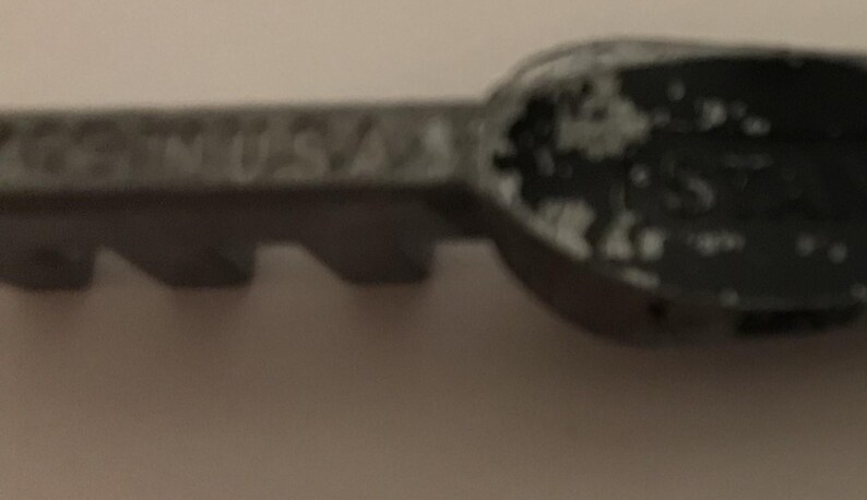 May include: Close-up of a gray metal key with the text "GS USA" embossed on the handle. The key has a worn, distressed appearance with areas of paint loss, revealing the metal beneath. The key's teeth are visible.