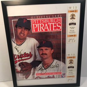 May include: A framed 1992 Pittsburgh Pirates magazine and scorecard featuring two baseball players, one holding a baseball. The magazine cover features the team logo and the text "OFFICIAL 1992 PITTSBURGH PIRATES" and "$2.00". The magazine also includes two game tickets with the text "GAME 21" and "LOUNGE BOX 390 A 12".