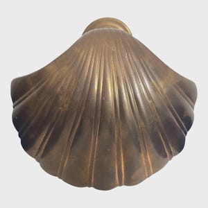 May include: A bronze-coloured, fan-shaped decorative object with a ridged surface. The object has a rounded top and a scalloped edge, resembling a seashell. The metallic finish gives it a vintage or antique appearance.