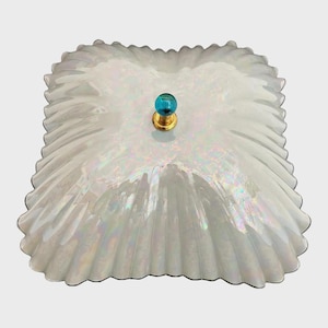 May include: A square, iridescent glass dish with a scalloped, ruffled edge and a glossy, pearlescent finish. The center features a small, round, turquoise-colored glass knob set on a gold-toned metal base, providing a distinct contrast.