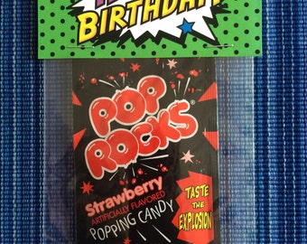 May include: A small, clear plastic bag with a green and black card attached to the top. The card says "Happy Birthday!" in a comic book style font. The bag contains a single serving of Pop Rocks candy, which is strawberry flavored. The candy package is black with red and white text that says "Pop Rocks", "Strawberry", "Artificially Flavored", "Popping Candy", "Taste the Explosion", and "Net Wt. 0.24 oz. (7g)".