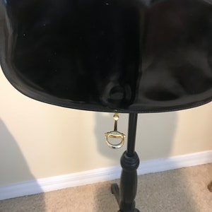 Pode incluir: Bolsa de m&atilde;o de couro patenteado preto com fecho de metal dourado pendurado em um suporte de madeira preto.