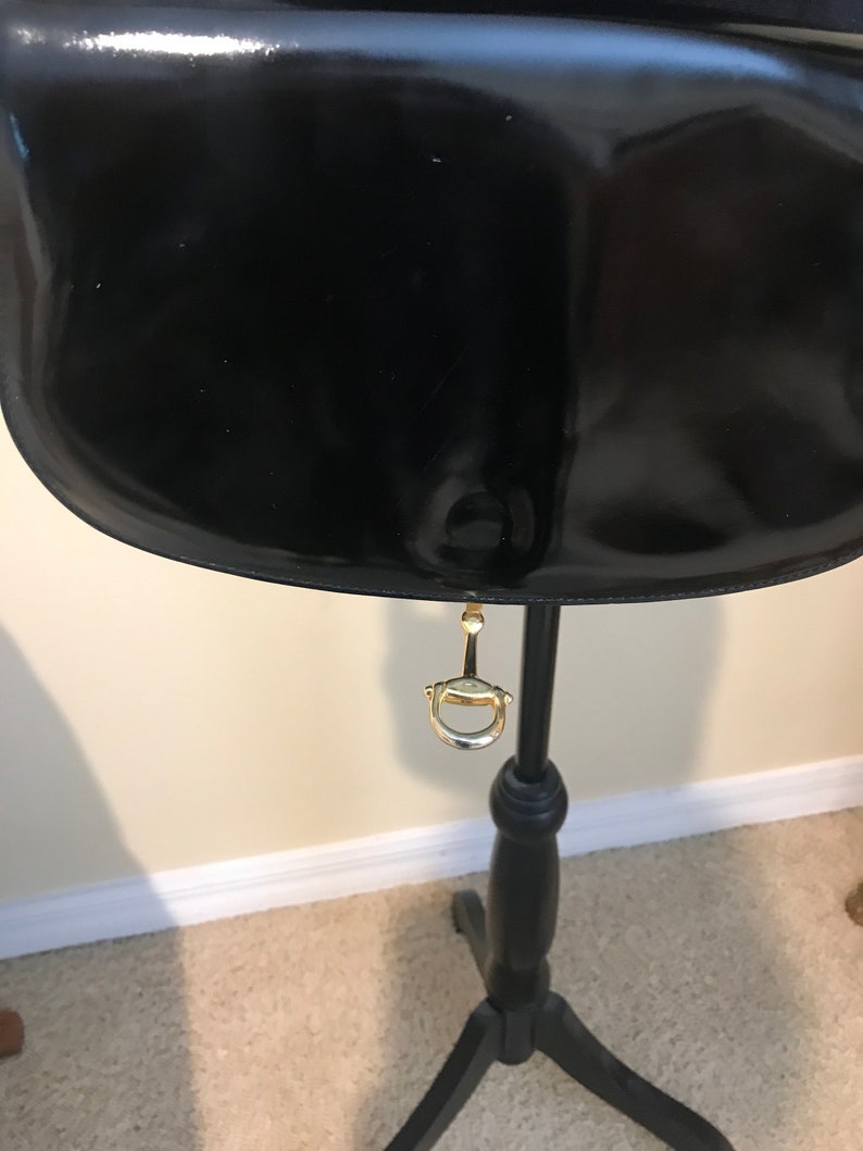 Pode incluir: Bolsa de m&atilde;o de couro patenteado preto com fecho de metal dourado pendurado em um suporte de madeira preto.