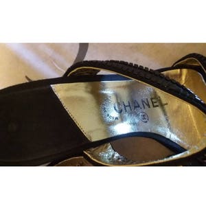 Pode incluir: Sand&aacute;lias de camur&ccedil;a preta com uma palmilha de couro dourado. A palmilha tem o texto "CHANEL" e um s&iacute;mbolo de marca registrada.