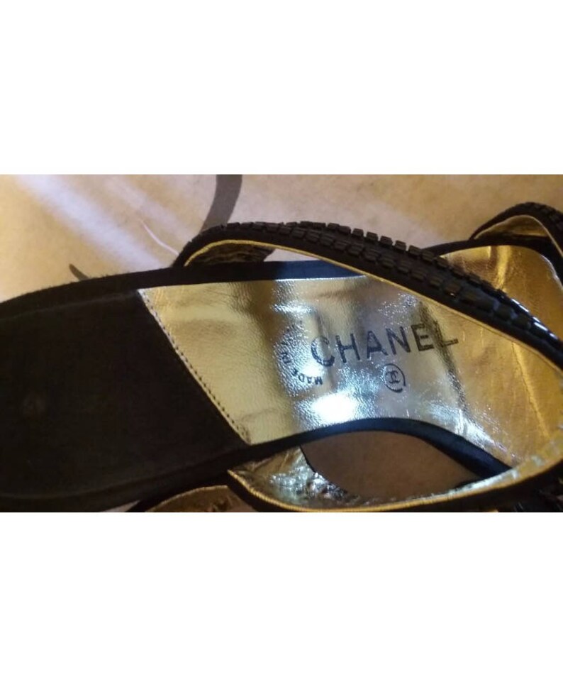 Pode incluir: Sand&aacute;lias de camur&ccedil;a preta com uma palmilha de couro dourado. A palmilha tem o texto "CHANEL" e um s&iacute;mbolo de marca registrada.