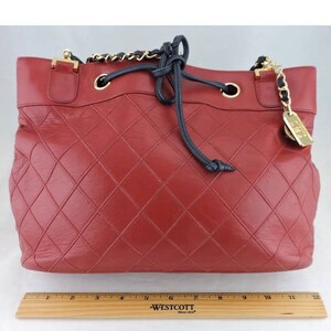 May include: A red quilted leather tote bag with a black drawstring closure and a gold chain strap. The bag has a gold metal tag with the word "Chanel" on it.