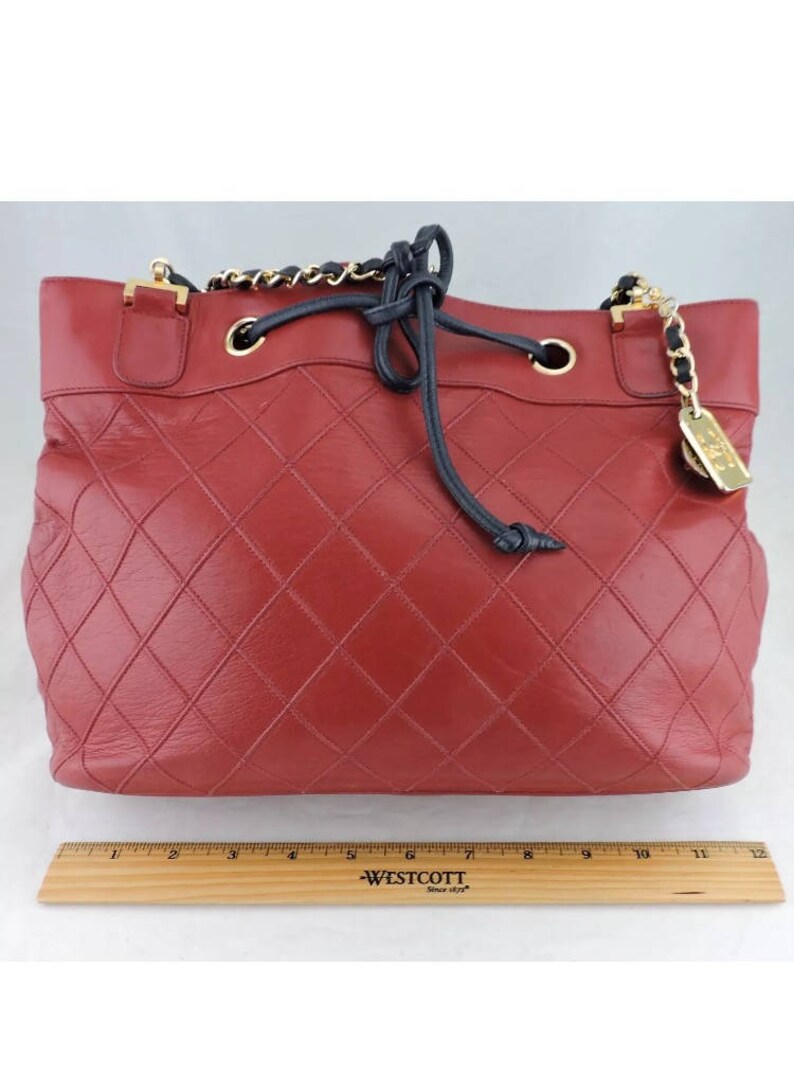 May include: A red quilted leather tote bag with a black drawstring closure and a gold chain strap. The bag has a gold metal tag with the word "Chanel" on it.
