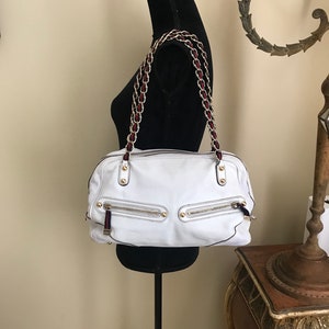 May include: White leather satchel handbag with a gold chain strap and red and black trim. The bag has multiple zippered pockets and gold accents.