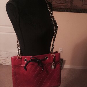 May include: A red quilted leather shoulder bag with a gold chain strap and a black drawstring closure. The bag has a gold logo on the front.