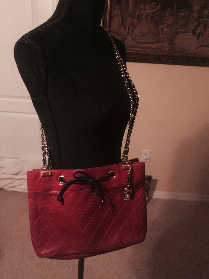 May include: A red quilted leather shoulder bag with a gold chain strap and a black drawstring closure. The bag has a gold logo on the front.