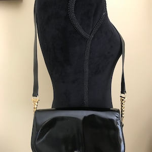 Pode incluir: Uma bolsa de ombro de couro patenteado preto com uma corrente de ouro e um fecho de ouro. A bolsa tem uma forma curva e um fecho de aba.