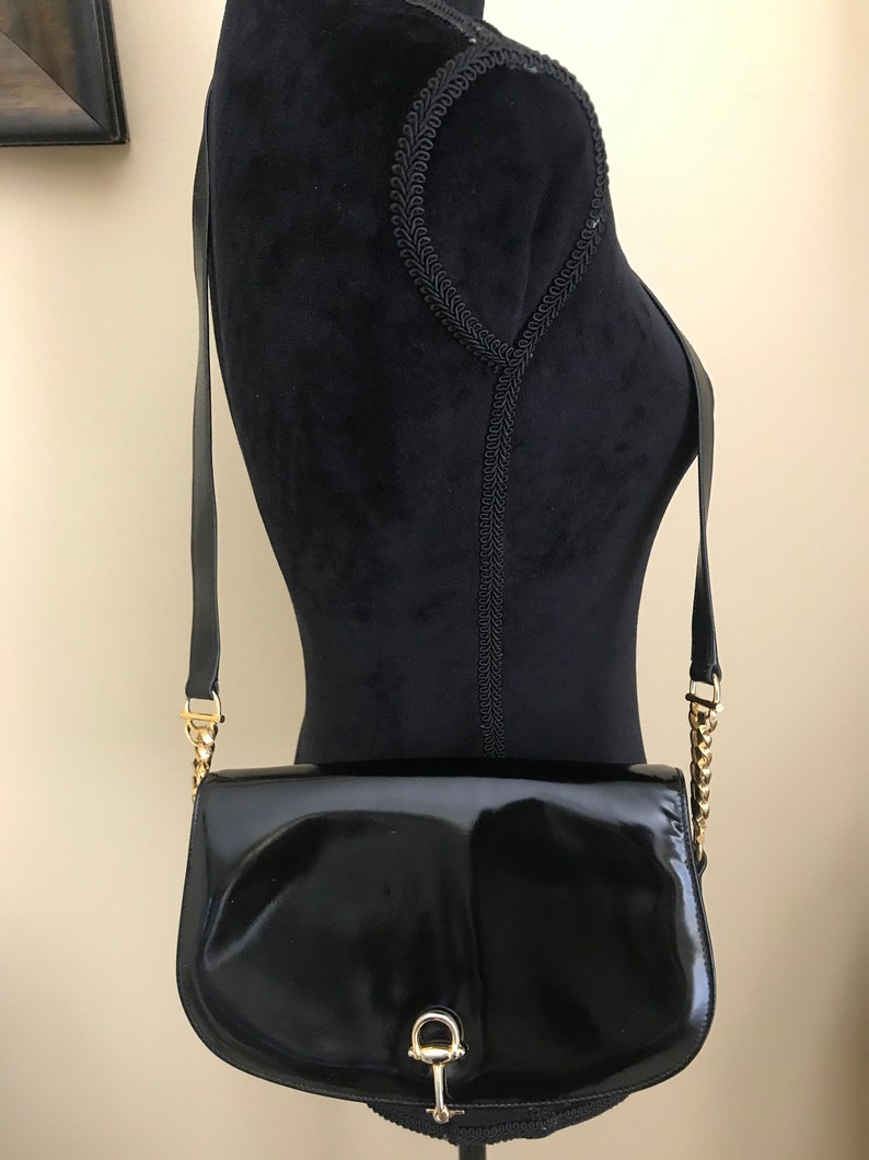 Pode incluir: Uma bolsa de ombro de couro patenteado preto com uma corrente de ouro e um fecho de ouro. A bolsa tem uma forma curva e um fecho de aba.