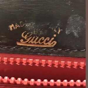 Pode incluir: Close-up de uma carteira de couro preta com um logotipo Gucci dourado e as palavras "Made in Italy". A carteira tem um z&iacute;per vermelho.