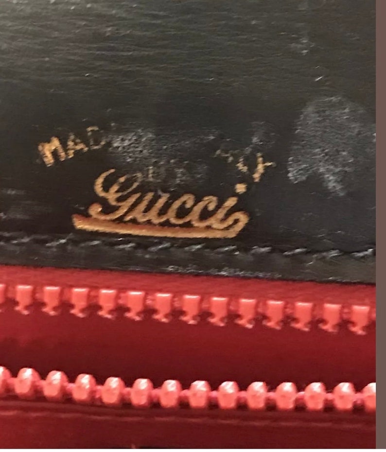 Pode incluir: Close-up de uma carteira de couro preta com um logotipo Gucci dourado e as palavras "Made in Italy". A carteira tem um z&iacute;per vermelho.
