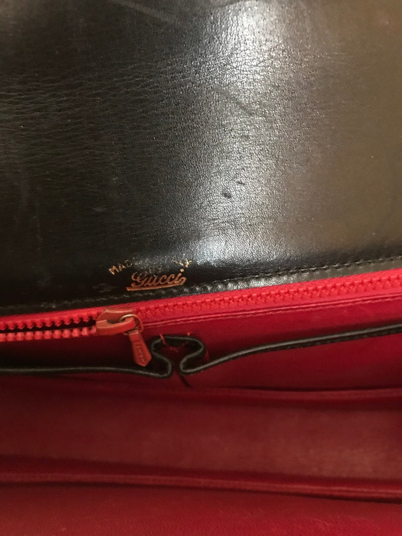 Pode incluir: O interior de uma bolsa de couro preta com forro vermelho. O z&iacute;per est&aacute; aberto e o forro &eacute; vis&iacute;vel. A bolsa tem uma etiqueta dourada que diz "Made in Italy" e "Gucci".