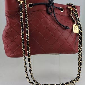 May include: A red quilted leather bucket bag with a black drawstring closure and a gold chain strap with black leather accents.