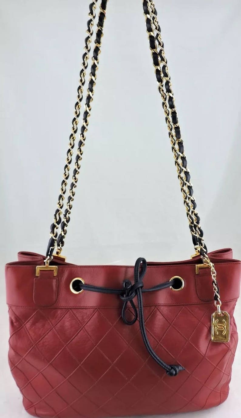 May include: A red quilted leather handbag with a black and gold chain strap and a drawstring closure. The bag has a gold Chanel logo charm.