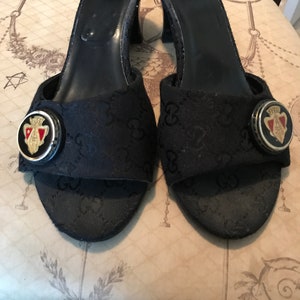 May include: A pair of black Gucci heeled sandals with a black and gold GG pattern. The sandals have a gold buckle with a red and green Gucci logo.