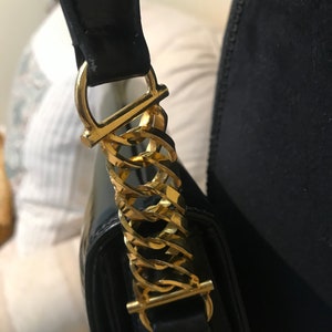 Pode incluir: Close-up de uma bolsa de m&atilde;o de couro preto com uma al&ccedil;a de corrente dourada. A al&ccedil;a tem uma fivela dourada e um la&ccedil;o dourado.