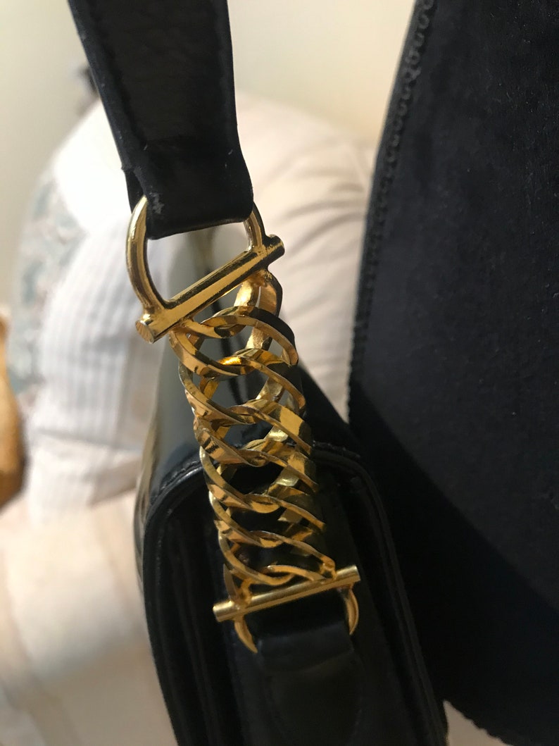 Pode incluir: Close-up de uma bolsa de m&atilde;o de couro preto com uma al&ccedil;a de corrente dourada. A al&ccedil;a tem uma fivela dourada e um la&ccedil;o dourado.