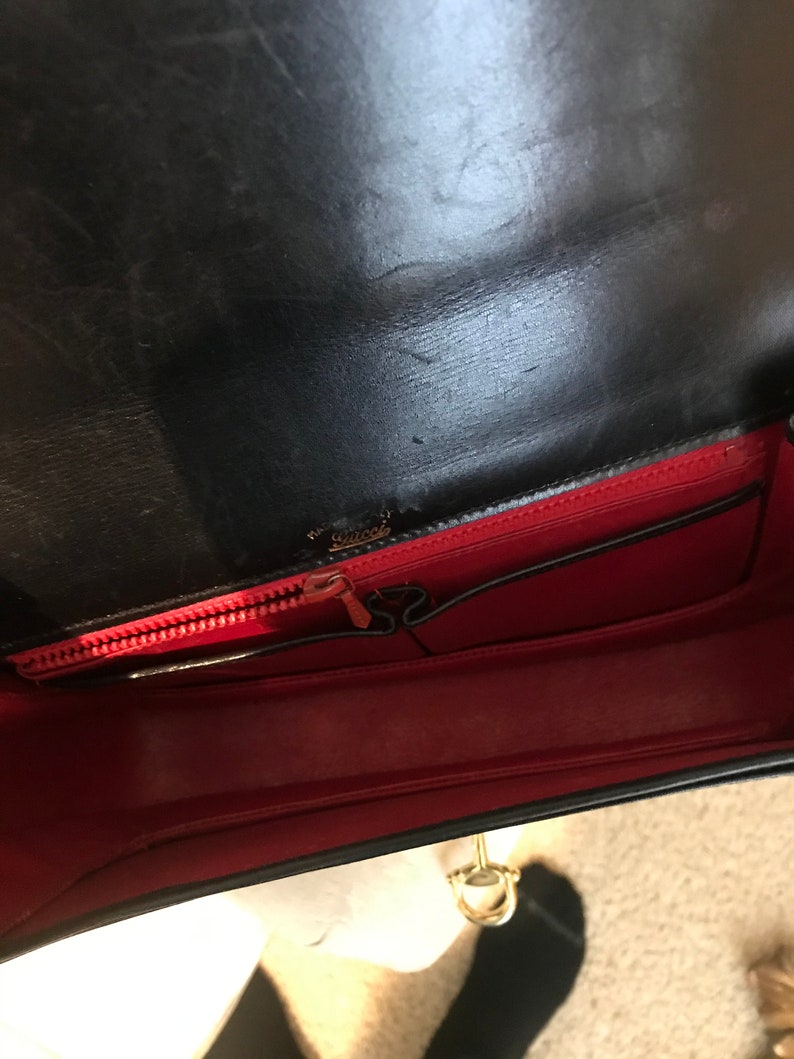 Pode incluir: O interior de uma bolsa de m&atilde;o de couro preto com forro vermelho. A bolsa tem fecho de correr e um fecho de metal dourado.