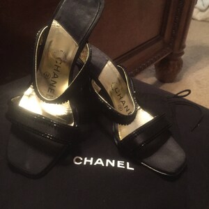 Pode incluir: Um par de sand&aacute;lias de salto alto de cetim preto com detalhes dourados e a palavra "CHANEL" na palmilha. As sand&aacute;lias est&atilde;o sobre uma bolsa de veludo preto com a palavra "CHANEL" impressa.